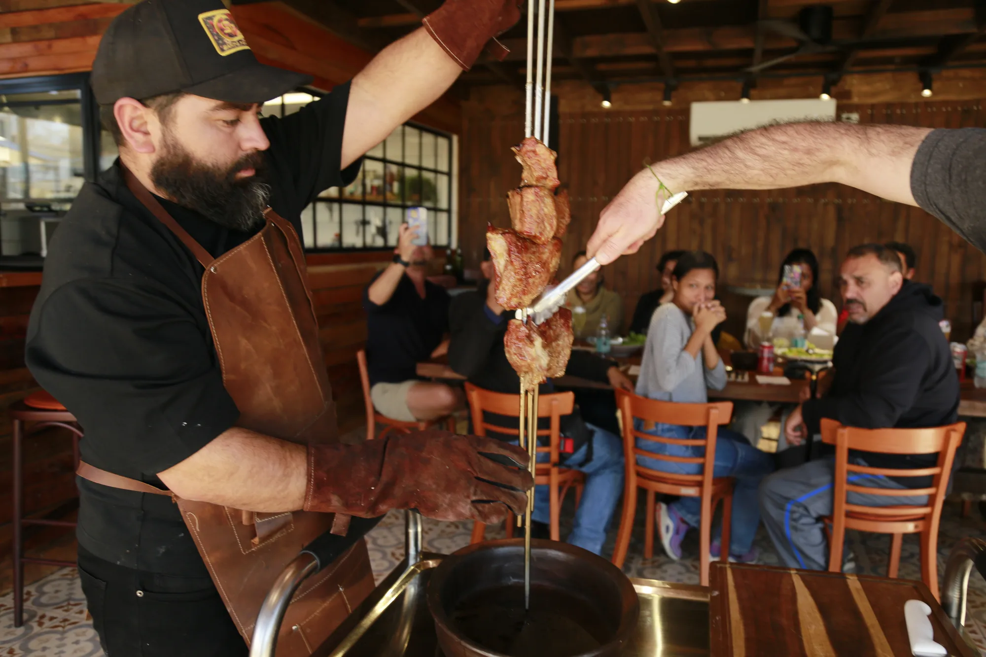 The chef lifting seared meat skewers from the fire, guests watching close by
